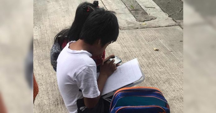 Irmãozinhos assistem às aulas na rua, sentados no chão, usando internet pública